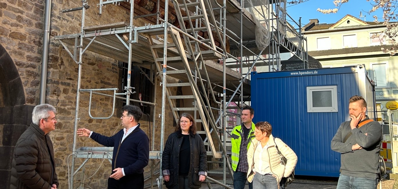 Mehrere Personen stehen bei einer Baustellenbegehung vor dem Alten Rathaus und betrachten die Arbeiten. An der historischen Steinfassade ist ein großes Gerüst mit Treppen aufgebaut. Eine Person erklärt gestikulierend Details zur Baustelle, während die anderen aufmerksam zuhören. Im Hintergrund steht ein blauer Baucontainer, daneben sind weitere Gebäude zu sehen. Sonniges Wetter mit klarem Himmel und blühenden Ästen am rechten Bildrand.