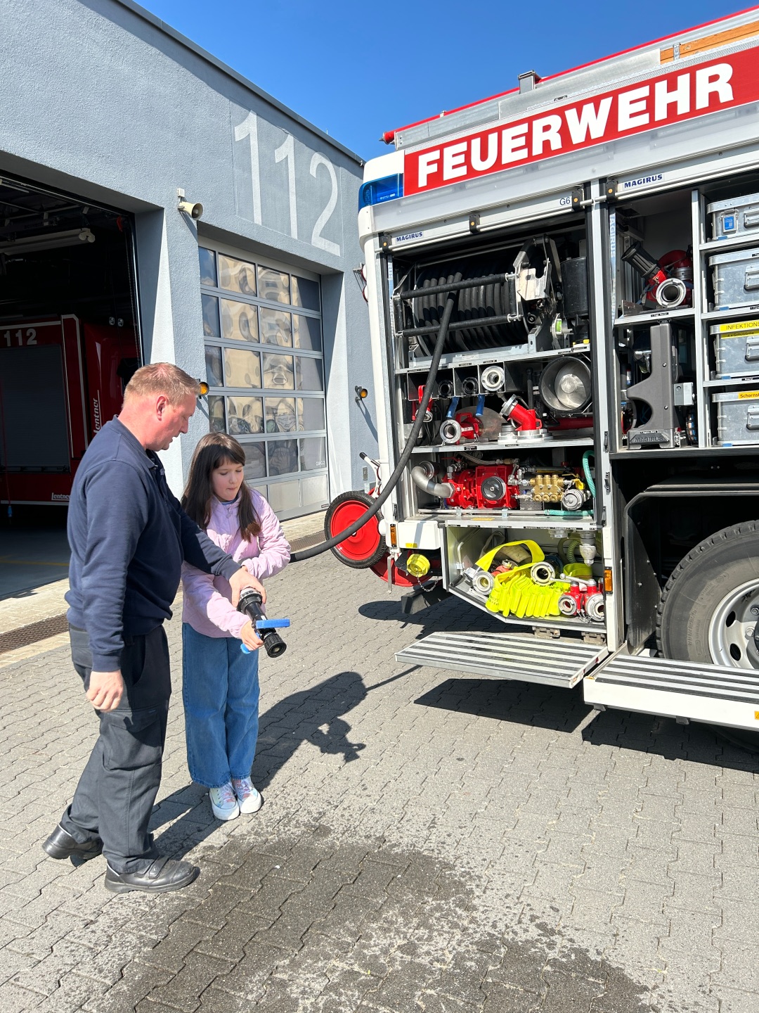 Ein Feuerwehrmann zeigt einem Mädchen vor einer Feuerwache, wie man einen Feuerwehrschlauch hält; neben ihnen steht ein geöffnetes Feuerwehrfahrzeug mit sichtbarer Ausrüstung.