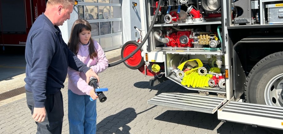 Ein Feuerwehrmann zeigt einem Mädchen vor einer Feuerwache, wie man einen Feuerwehrschlauch hält; neben ihnen steht ein geöffnetes Feuerwehrfahrzeug mit sichtbarer Ausrüstung.