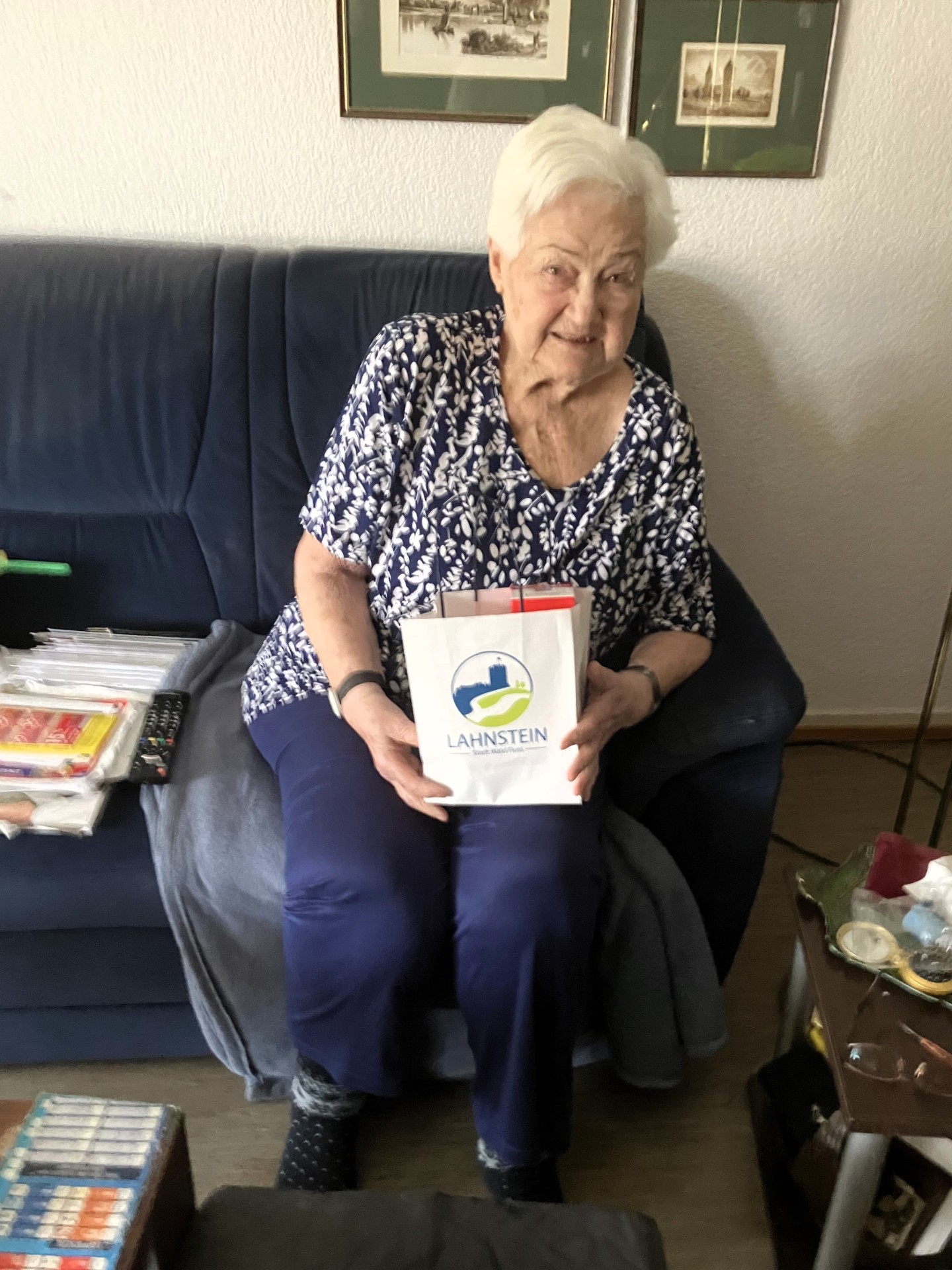 Eine ältere Frau sitzt auf einem Sofa und hält eine Geschenktüte mit der Aufschrift „Lahnstein“ in den Händen. Sie lächelt freundlich in die Kamera. Neben ihr liegen Zeitschriften und eine Fernbedienung, im Hintergrund hängen gerahmte Bilder an der Wand.