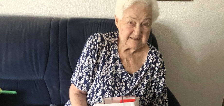 Sigrid Paulus feierte ihren 90. Geburtstag in Lahnstein Eine ältere Frau sitzt auf einem Sofa und hält eine Geschenktüte mit der Aufschrift „Lahnstein“ in den Händen. Sie lächelt freundlich in die Kamera. Neben ihr liegen Zeitschriften und eine Fernbedienung, im Hintergrund hängen gerahmte Bilder an der Wand.