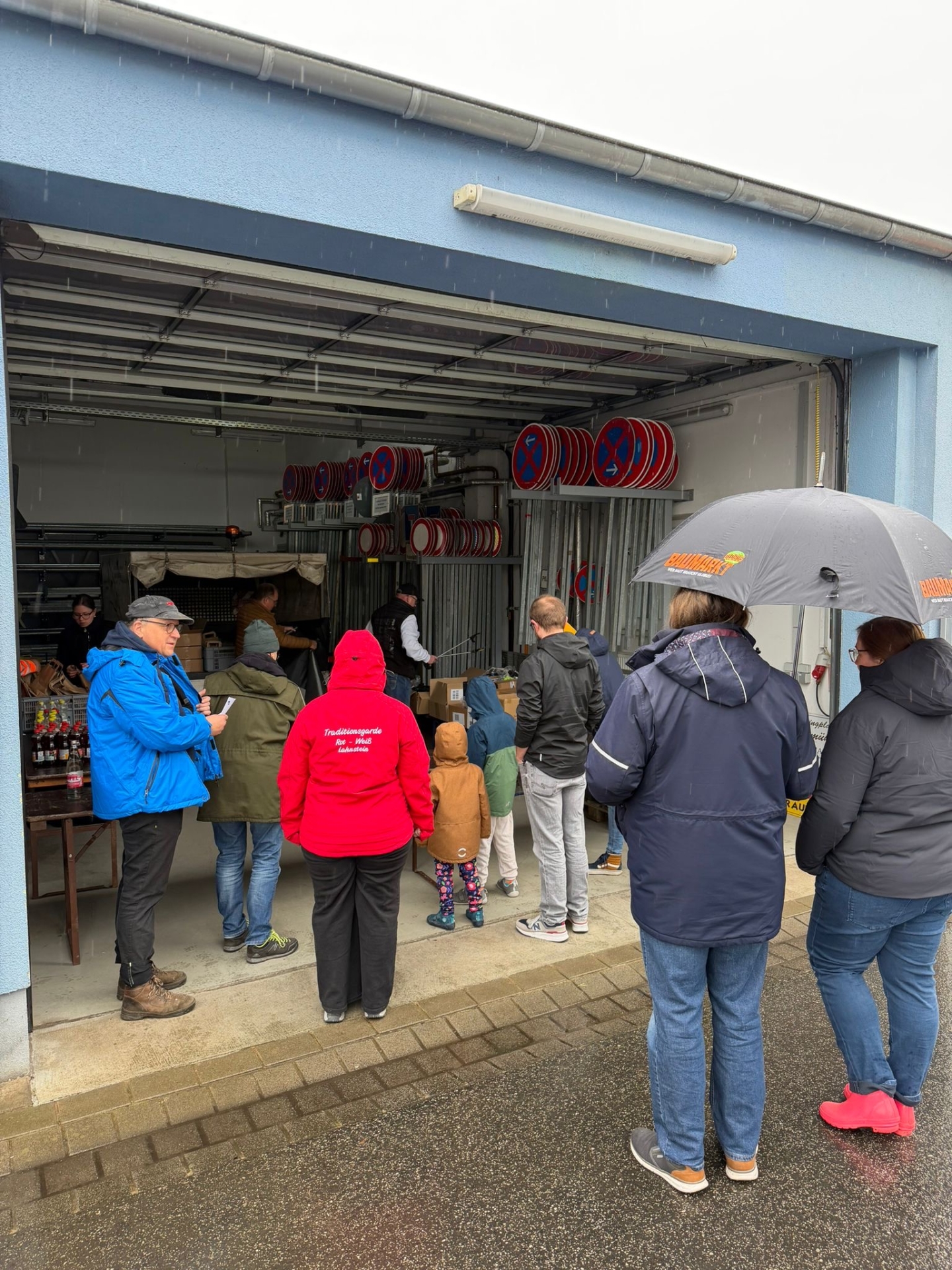 Eine Gruppe von Menschen steht bei regnerischem Wetter vor und in einer offenen Garage oder Lagerhalle. Sie tragen wetterfeste Kleidung; eine Person hält einen Regenschirm. Im Hintergrund sind zahlreiche Verkehrsschilder (Halteverbot) ordentlich in Regalen gelagert. Es scheint sich um die Ausgabe von Material oder eine Lagebesprechung für eine gemeinschaftliche Aktion zu handeln.