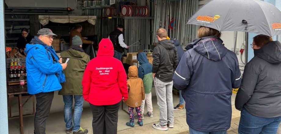 Eine Gruppe von Menschen steht bei regnerischem Wetter vor und in einer offenen Garage oder Lagerhalle. Sie tragen wetterfeste Kleidung; eine Person hält einen Regenschirm. Im Hintergrund sind zahlreiche Verkehrsschilder (Halteverbot) ordentlich in Regalen gelagert. Es scheint sich um die Ausgabe von Material oder eine Lagebesprechung für eine gemeinschaftliche Aktion zu handeln.