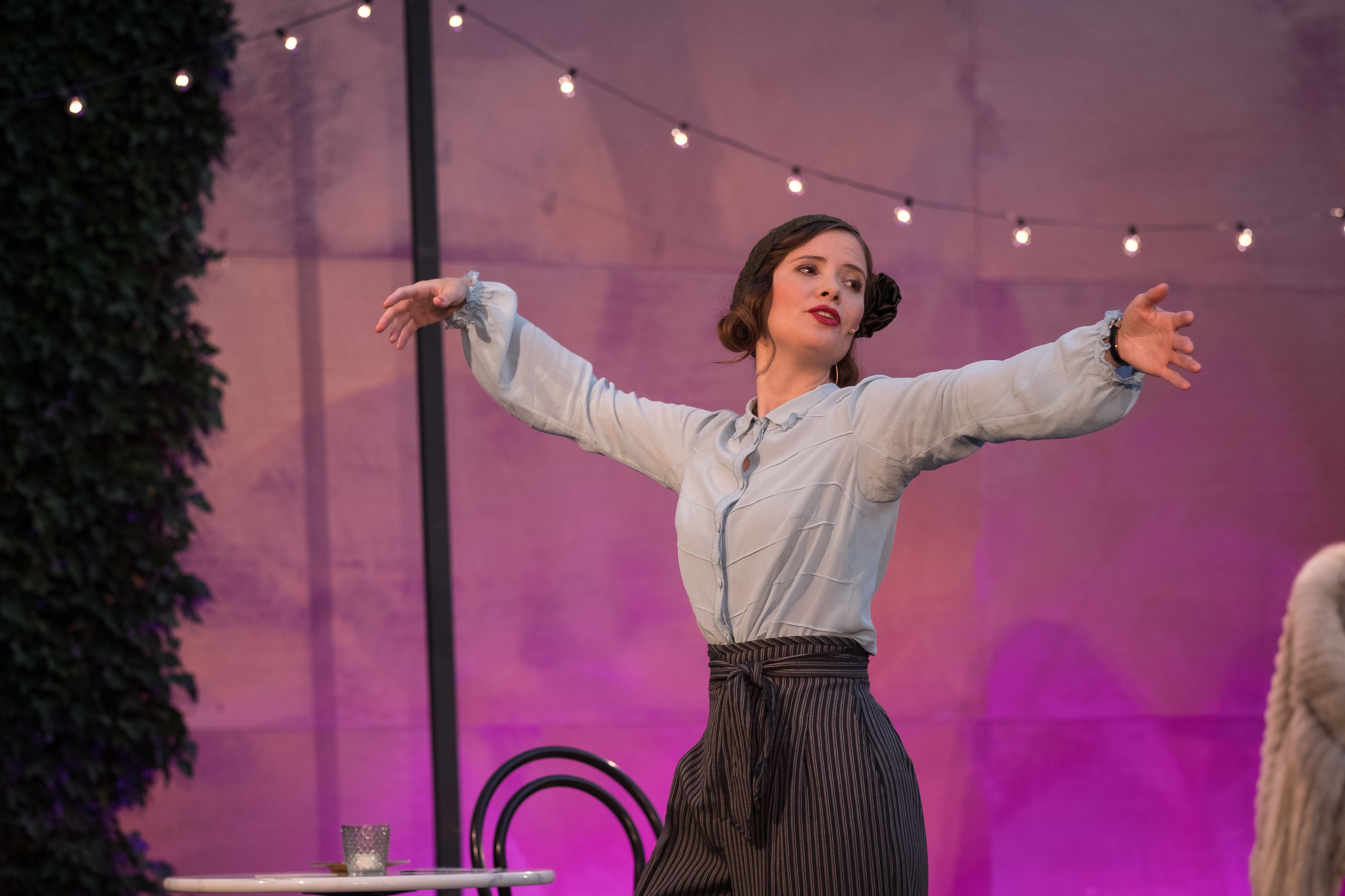 Eine Darstellerin steht mit weit ausgestreckten Armen auf einer Theaterbühne. Sie trägt eine helle Bluse und eine dunkle, weit geschnittene Hose. Der Hintergrund ist violett beleuchtet und mit einer Lichterkette dekoriert.