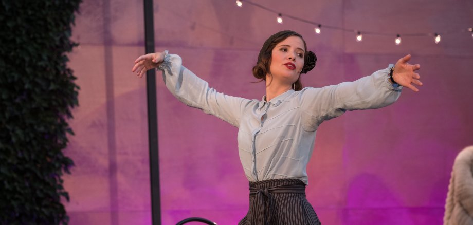 Das kunstseidene Mädchen Eine Darstellerin steht mit weit ausgestreckten Armen auf einer Theaterbühne. Sie trägt eine helle Bluse und eine dunkle, weit geschnittene Hose. Der Hintergrund ist violett beleuchtet und mit einer Lichterkette dekoriert.