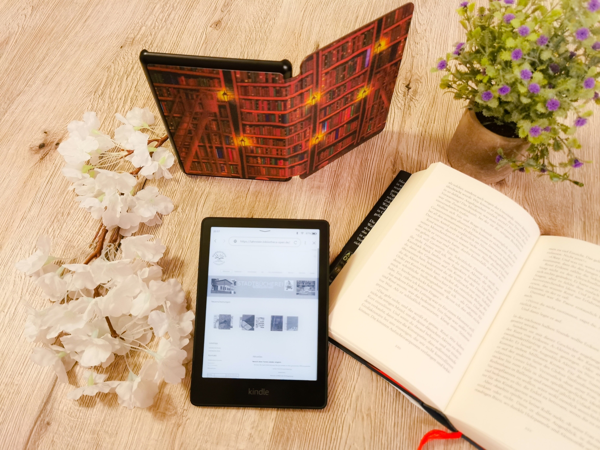 Ein eBook-Reader liegt neben einem aufgeschlagenem Buch auf einem Tisch mit Blumendeko.