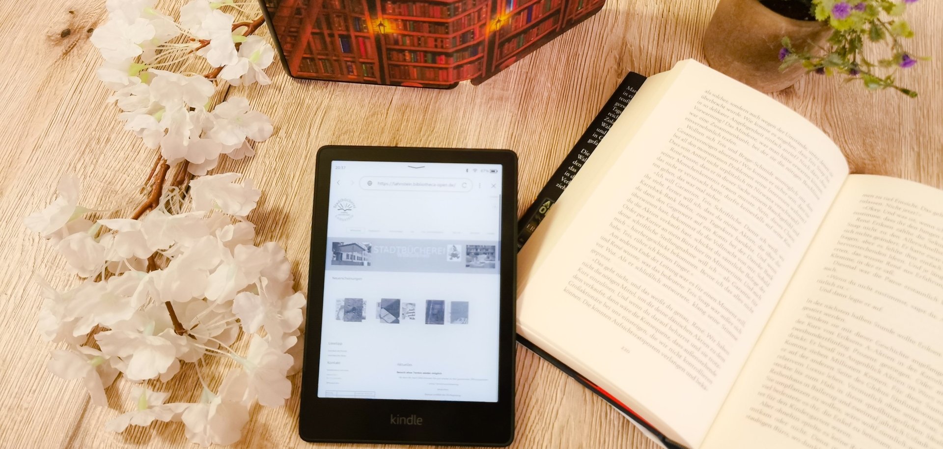 Ein eBook-Reader liegt neben einem aufgeschlagenem Buch auf einem Tisch mit Blumendeko.
