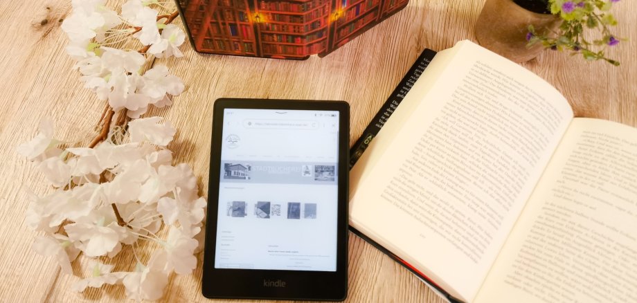 eBook-Sprechstunden in der Stadtbücherei Lahnstein Ein eBook-Reader liegt neben einem aufgeschlagenem Buch auf einem Tisch mit Blumendeko.