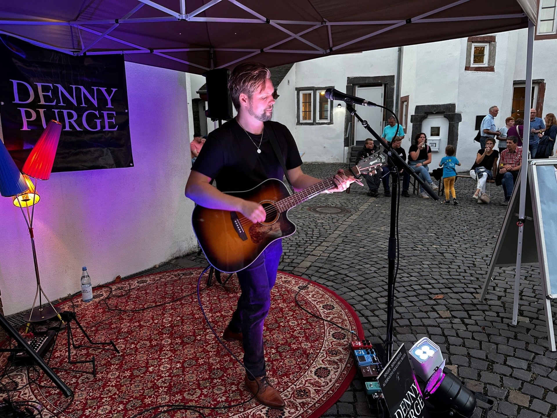 Ein Musiker steht unter einem Pavillon und spielt eine Akustikgitarre vor einem Mikrofon. Er trägt ein schwarzes T-Shirt und Jeans und steht auf einem gemusterten Teppich. Neben ihm sind Lautsprecher und Bühnenequipment aufgebaut. Im Hintergrund sitzen und stehen einige Zuschauer in einem gepflasterten Innenhof vor weißen Gebäuden und beobachten die kleine Live-Performance.
