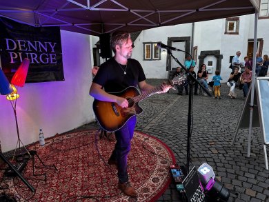 Ein Musiker steht unter einem Pavillon und spielt eine Akustikgitarre vor einem Mikrofon. Er trägt ein schwarzes T-Shirt und Jeans und steht auf einem gemusterten Teppich. Neben ihm sind Lautsprecher und Bühnenequipment aufgebaut. Im Hintergrund sitzen und stehen einige Zuschauer in einem gepflasterten Innenhof vor weißen Gebäuden und beobachten die kleine Live-Performance.