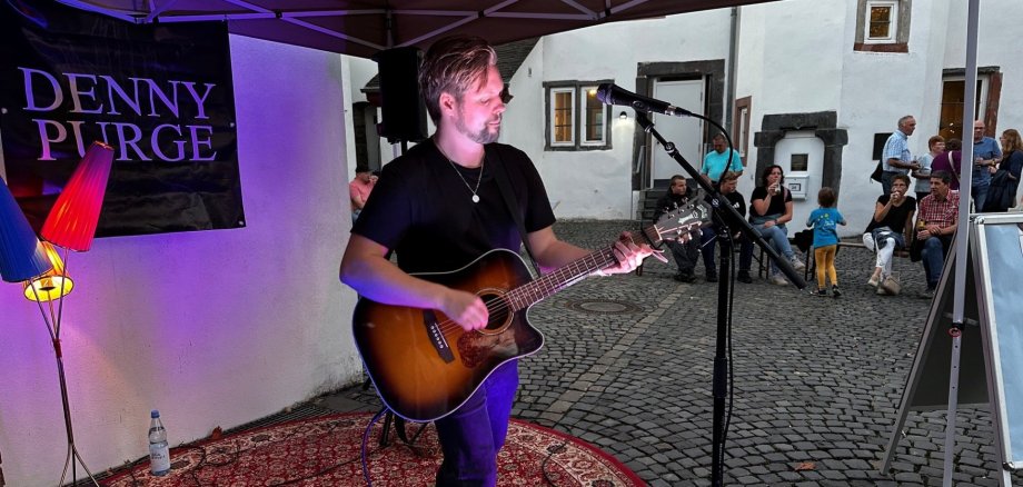 Lahnsteiner Treff Ein Musiker steht unter einem Pavillon und spielt eine Akustikgitarre vor einem Mikrofon. Er trägt ein schwarzes T-Shirt und Jeans und steht auf einem gemusterten Teppich. Neben ihm sind Lautsprecher und Bühnenequipment aufgebaut. Im Hintergrund sitzen und stehen einige Zuschauer in einem gepflasterten Innenhof vor weißen Gebäuden und beobachten die kleine Live-Performance.