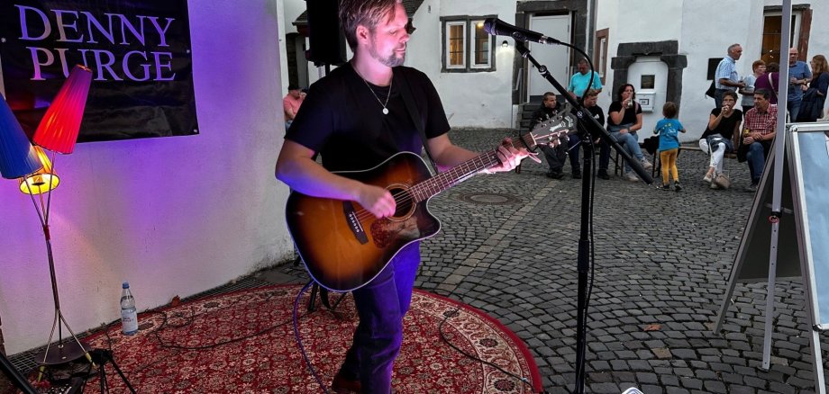 Ein Musiker steht unter einem Pavillon und spielt eine Akustikgitarre vor einem Mikrofon. Er trägt ein schwarzes T-Shirt und Jeans und steht auf einem gemusterten Teppich. Neben ihm sind Lautsprecher und Bühnenequipment aufgebaut. Im Hintergrund sitzen und stehen einige Zuschauer in einem gepflasterten Innenhof vor weißen Gebäuden und beobachten die kleine Live-Performance.