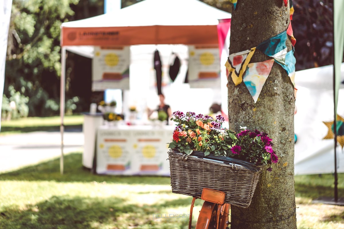 Ein Fahrrad mit geflochtenem Korb voller bunter Blumen lehnt an einem Baum, der mit kleinen, farbigen Wimpeln geschmückt ist. Im Hintergrund steht ein Pavillon mit Infostand auf einer grünen Wiese, der eine gemütliche, festivalartige Atmosphäre vermittelt.