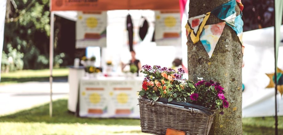 Ein Fahrrad mit geflochtenem Korb voller bunter Blumen lehnt an einem Baum, der mit kleinen, farbigen Wimpeln geschmückt ist. Im Hintergrund steht ein Pavillon mit Infostand auf einer grünen Wiese, der eine gemütliche, festivalartige Atmosphäre vermittelt.