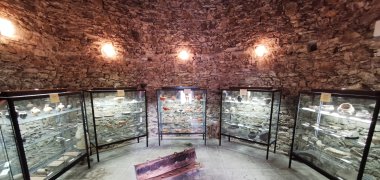 Runder Ausstellungsraum mit Steinwänden und mehreren Glasvitrinen, in denen archäologische Funde wie Keramik und Werkzeuge präsentiert werden.