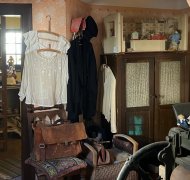 Historisch eingerichteter Wohnraum mit alten Möbeln, Kleidung und Haushaltsgegenständen; im Vordergrund eine mechanische Wäschemangel, im Hintergrund ein Raum mit Puppen und Alltagsobjekten.