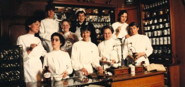 Historische Apothekenszene mit mehreren Personen in weißen Kitteln hinter einem Verkaufstresen, umgeben von Gefäßen, Waagen und Medikamenten.