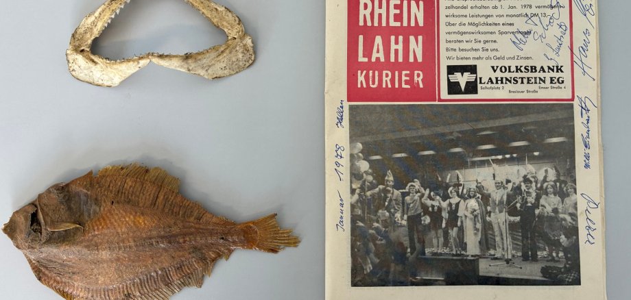 Auf einer hellen Fläche liegen drei Gegenstände: rechts eine Ausgabe des ‚Rhein-Lahn-Kuriers‘ von 1978 mit handschriftlichen Unterschriften, links oben ein gebogener Fischkiefer mit Zähnen und links unten ein getrockneter, halbierter Fisch.