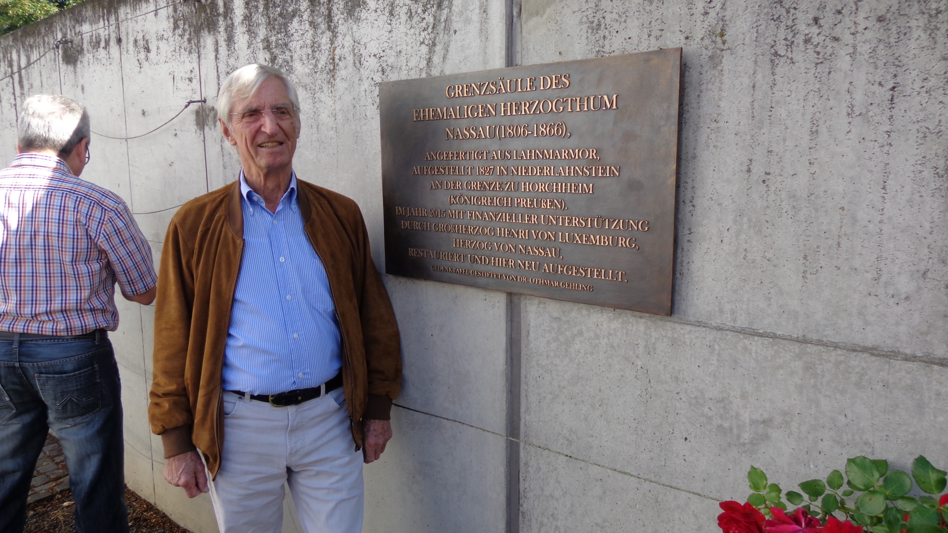 Ein älterer Mann steht neben einer Bronzetafel zur Grenzsäule des Herzogtums Nassau an einer Betonwand im Freien. Im Hintergrund ist ein weiterer Mann von hinten zu sehen.