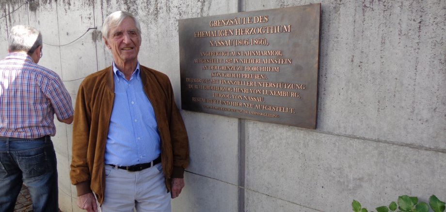 Ein älterer Mann steht neben einer Bronzetafel zur Grenzsäule des Herzogtums Nassau an einer Betonwand im Freien. Im Hintergrund ist ein weiterer Mann von hinten zu sehen.