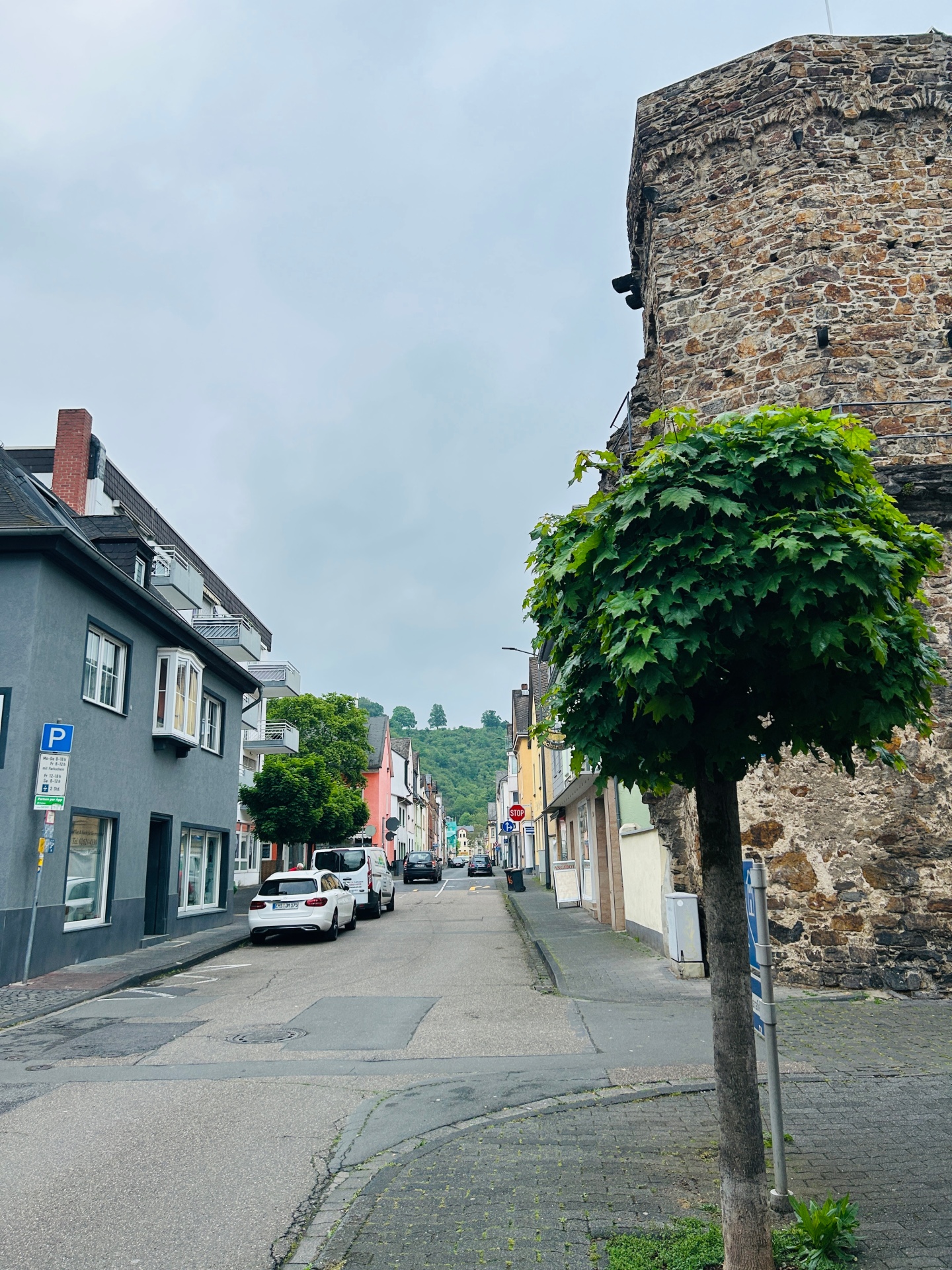 Schmale Wohnstraße mit parkenden Autos und bunten Häusern, die leicht bergauf zu einer grünen Hügellandschaft führt. Im Vordergrund steht ein rund geschnittener Baum neben einer alten, runden Steinmauer unter grauem, bewölktem Himmel.