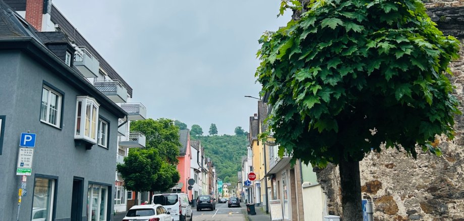 Schmale Wohnstraße mit parkenden Autos und bunten Häusern, die leicht bergauf zu einer grünen Hügellandschaft führt. Im Vordergrund steht ein rund geschnittener Baum neben einer alten, runden Steinmauer unter grauem, bewölktem Himmel.