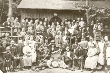 Historische Schwarz-Weiß-Aufnahme aus dem Jahr 1925. Eine große Gruppe von Menschen in zeitgenössischer Kleidung – Männer in Anzügen, Frauen in langen Kleidern und Schürzen sowie Kinder – posiert vor einer rustikalen Holzhalle mit verzierter Fassade. Im Vordergrund sitzt eine Kapelle mit Blasinstrumenten und Trommeln.