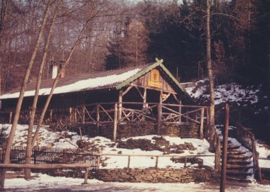 Farbfotografie aus dem Jahr 1970. Zu sehen ist eine rustikale, bewirtschaftete Waldschenke mit Satteldach und einer hölzernen Terrasse auf einem Steinfundament. Das Gebäude liegt an einem Hang im Wald, der Boden ist leicht mit Schnee bedeckt. Ein Schild über dem Eingang weist auf die Bewirtung hin.
