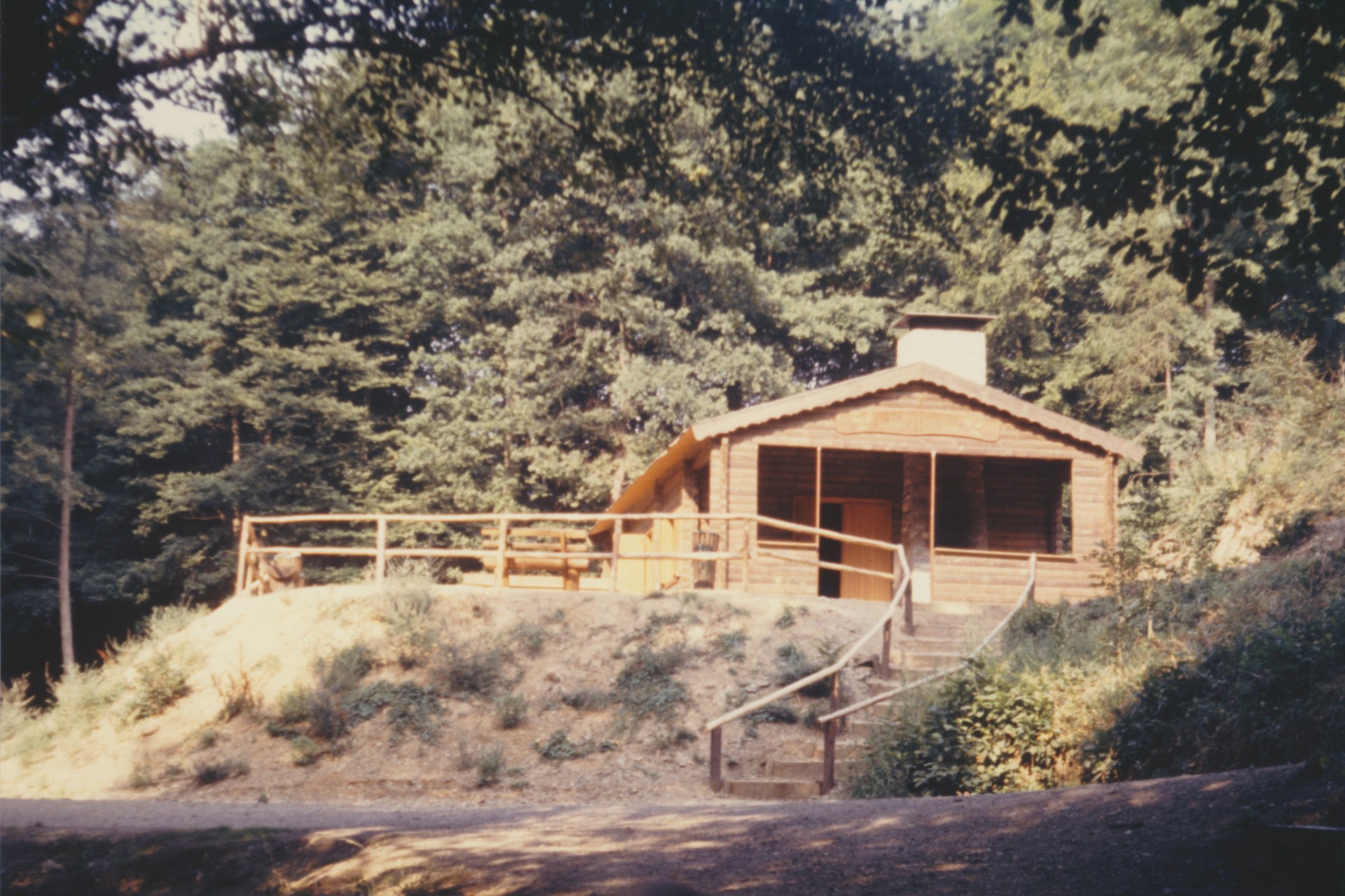 Farbfotografie aus der Entstehungszeit der aktuellen Hütte in der Ruppertsklamm aus dem Jahr 1976. Die neue, offene Blockhütte aus hellem Holz steht auf einer Anhöhe im Wald. Sie verfügt über ein flaches Satteldach mit Schornstein und eine davorliegende Terrasse mit Holzgeländer. Eine Steintreppe führt vom Wanderweg hinauf zur Hütte.