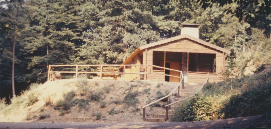 Farbfotografie aus der Entstehungszeit der aktuellen Hütte in der Ruppertsklamm aus dem Jahr 1976. Die neue, offene Blockhütte aus hellem Holz steht auf einer Anhöhe im Wald. Sie verfügt über ein flaches Satteldach mit Schornstein und eine davorliegende Terrasse mit Holzgeländer. Eine Steintreppe führt vom Wanderweg hinauf zur Hütte.