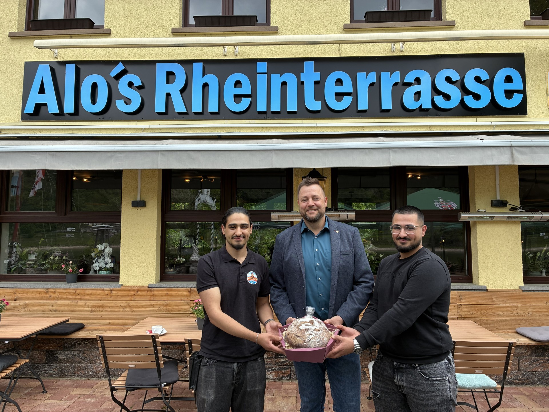 Gruppenfoto vor dem neu eröffneten Restaurant „Alo‘s Rheinterrasse“ in Niederlahnstein. Zu sehen sind die beiden Betreiber Robin und Rodi Hussein gemeinsam mit Oberbürgermeister Lennart Siefert. Im Hintergrund sind die Außenterrasse des Restaurants sowie der Rhein zu erkennen.