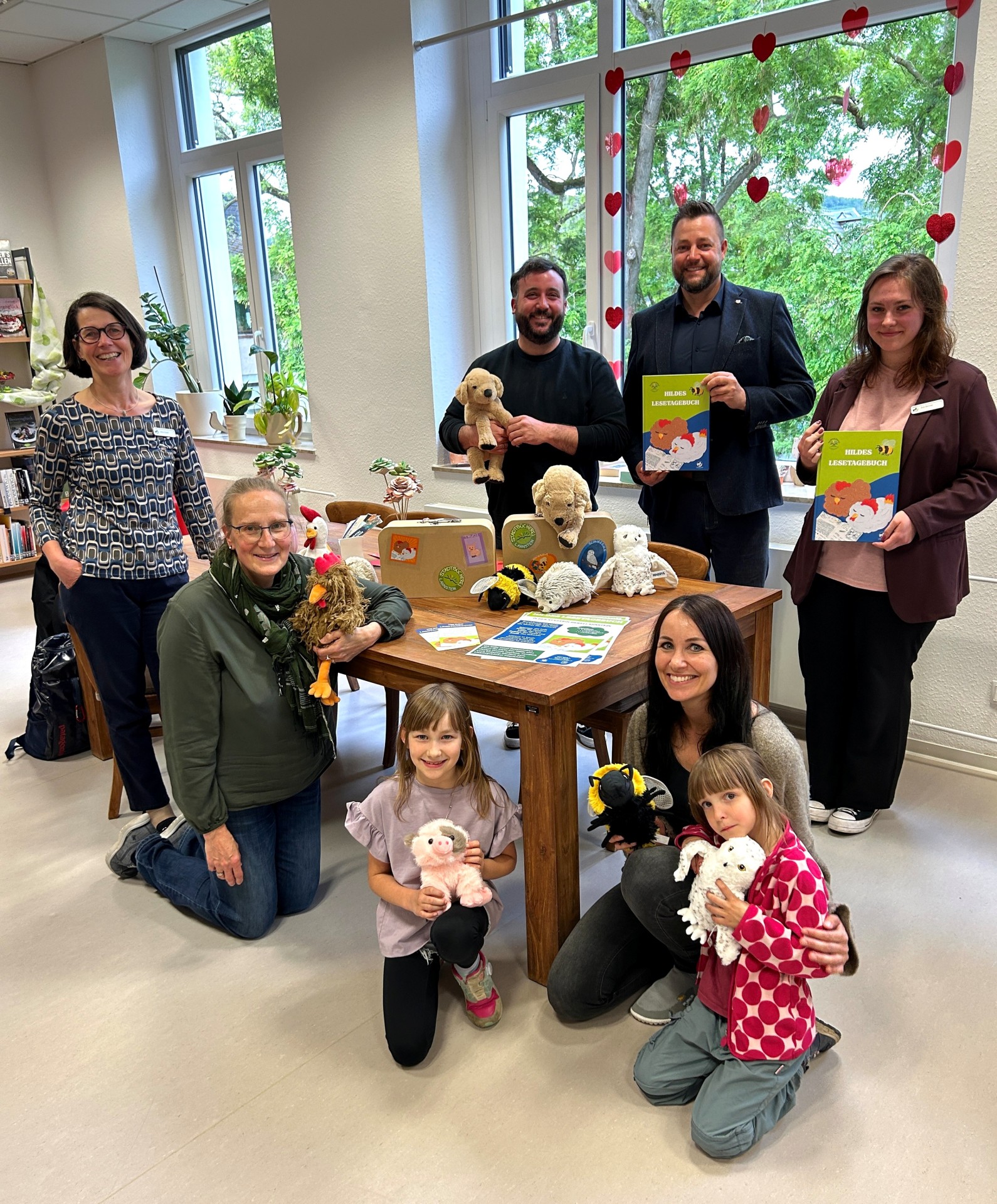 Gruppenfoto in der Stadtbücherei Lahnstein zur Vorstellung des Projekts „Hildes Lesetagebuch“. Vier Erwachsene stehen im Hintergrund, zwei Frauen knien im Vordergrund neben zwei Grundschulkindern um einen Holztisch herum. Sie halten bunte Lesetagebücher, zwei kleine Koffer mit Aufklebern sowie verschiedene Plüschtiere – darunter einen Hund, ein Huhn, ein Schwein und eine Biene – lächelnd in die Kamera. Im Hintergrund sind helle Fenster mit einer Herzchen-Dekoration und Buchregale zu sehen.