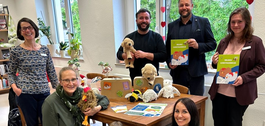 Stadtbücherei Lahnstein startet „Hildes Lesetagebuch“ Gruppenfoto in der Stadtbücherei Lahnstein zur Vorstellung des Projekts „Hildes Lesetagebuch“. Vier Erwachsene stehen im Hintergrund, zwei Frauen knien im Vordergrund neben zwei Grundschulkindern um einen Holztisch herum. Sie halten bunte Lesetagebücher, zwei kleine Koffer mit Aufklebern sowie verschiedene Plüschtiere – darunter einen Hund, ein Huhn, ein Schwein und eine Biene – lächelnd in die Kamera. Im Hintergrund sind helle Fenster mit einer Herzchen-Dekoration und Buchregale zu sehen.