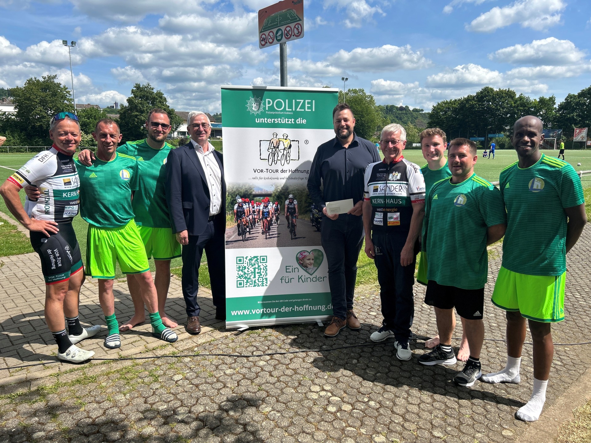 Eine Gruppe von Männern steht auf einem asphaltierten Platz vor einem großen Informationsbanner der „Vor-Tour der Hoffnung“, die sich für Kinder einsetzt. Einige Männer tragen Fußballtrikots, andere tragen Radsportkleidung oder formelle Kleidung. Ein Mann in der Mitte hält einen Umschlag in der Hand. Im Hintergrund sind ein Fußballfeld und blauer Himmel zu sehen.