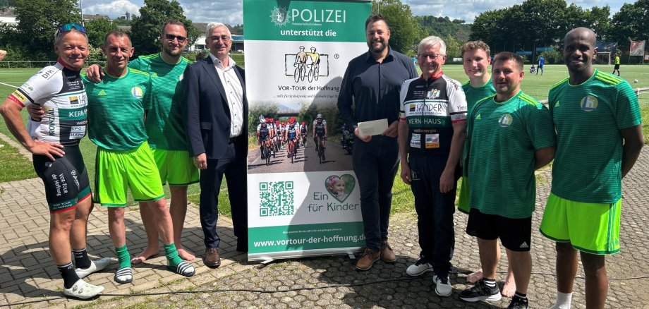 Benefiz-Fußballturnier für den guten Zweck in Lahnstein Eine Gruppe von Männern steht auf einem asphaltierten Platz vor einem großen Informationsbanner der „Vor-Tour der Hoffnung“, die sich für Kinder einsetzt. Einige Männer tragen Fußballtrikots, andere tragen Radsportkleidung oder formelle Kleidung. Ein Mann in der Mitte hält einen Umschlag in der Hand. Im Hintergrund sind ein Fußballfeld und blauer Himmel zu sehen.