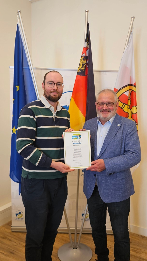 Zwei Männer stehen vor Europa-, Deutschland- und Stadtflagge und halten gemeinsam die Urkunde zur erneuten Auszeichnung Lahnsteins als Fairtrade-Stadt in die Kamera. Links steht ein jüngerer Mann im gestreiften Pullover, rechts Bürgermeister Johannes Lauer im blauen Sakko.