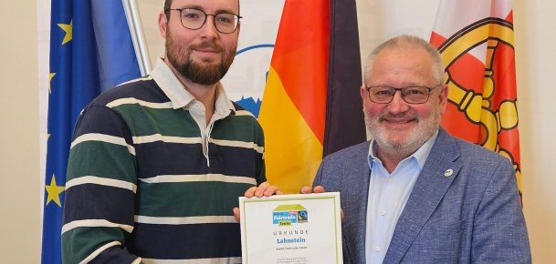 Zwei Männer stehen vor Europa-, Deutschland- und Stadtflagge und halten gemeinsam die Urkunde zur erneuten Auszeichnung Lahnsteins als Fairtrade-Stadt in die Kamera. Links steht ein jüngerer Mann im gestreiften Pullover, rechts Bürgermeister Johannes Lauer im blauen Sakko.