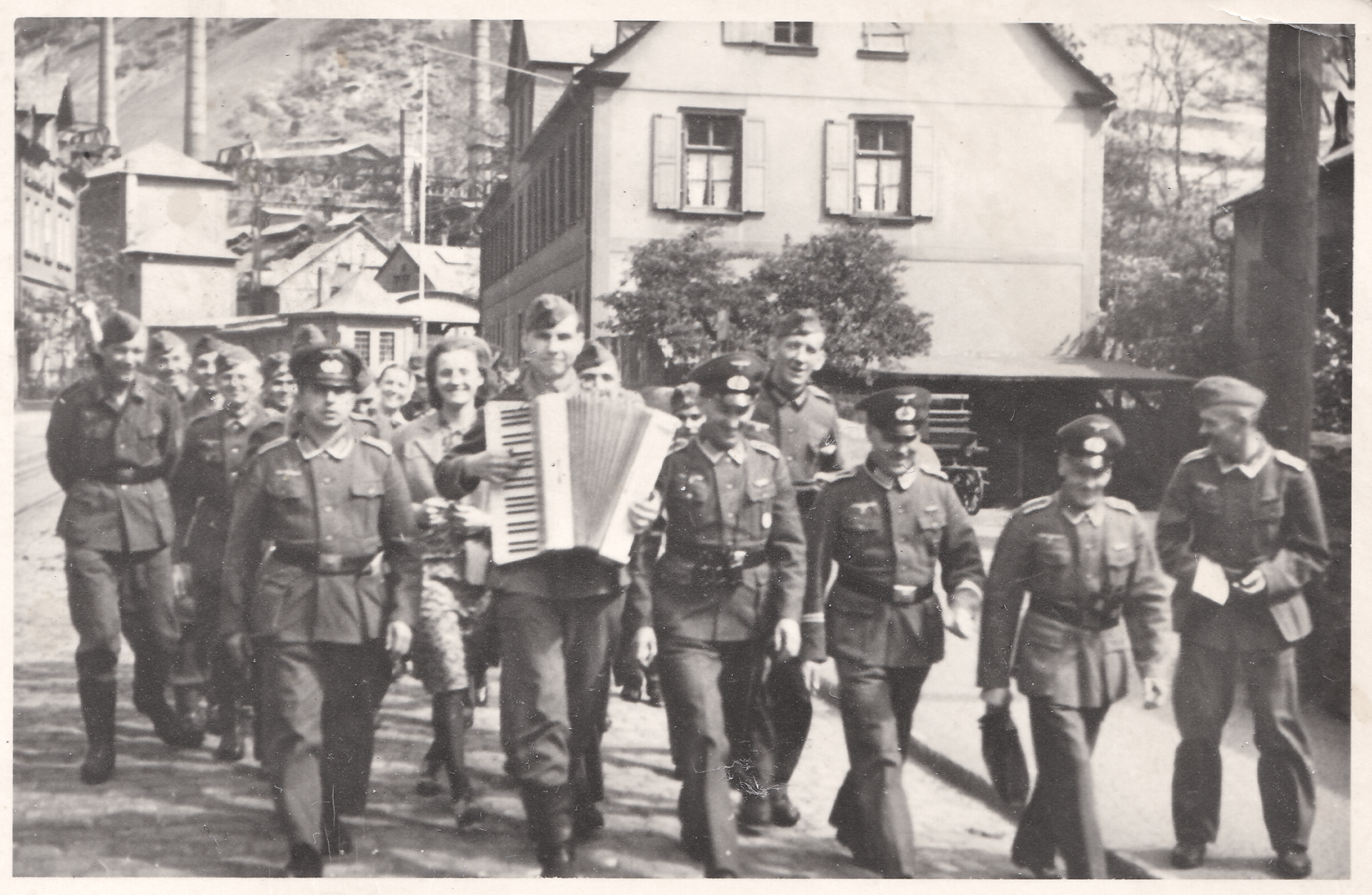 Schwarz-Weiß-Bild einer Gruppe Soldaten in Uniform auf einer Straße. Ein Mann in erster Reihe spielt ein Akkordeon.