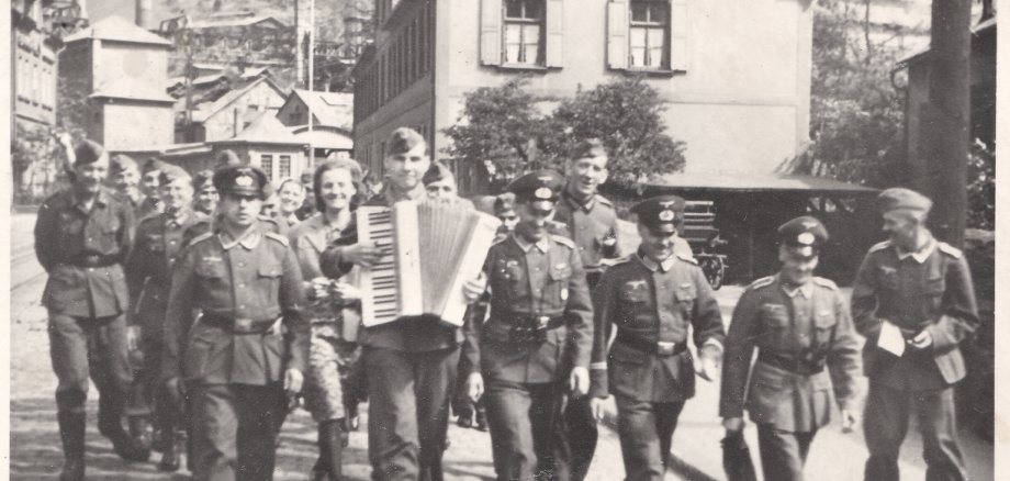 Schwarz-Weiß-Bild einer Gruppe Soldaten in Uniform auf einer Straße. Ein Mann in erster Reihe spielt ein Akkordeon.