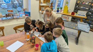 Mehrere Kinder sitzen gemeinsam mit einer Frau an einem großen Holztisch in der Stadtbücherei Lahnstein und bearbeiten konzentriert Arbeitsblätter. Auf dem Tisch stehen Getränke und Bastelmaterialien, im Hintergrund sind Bücherregale, Fensterdekorationen und ein Puzzlebereich zu sehen.