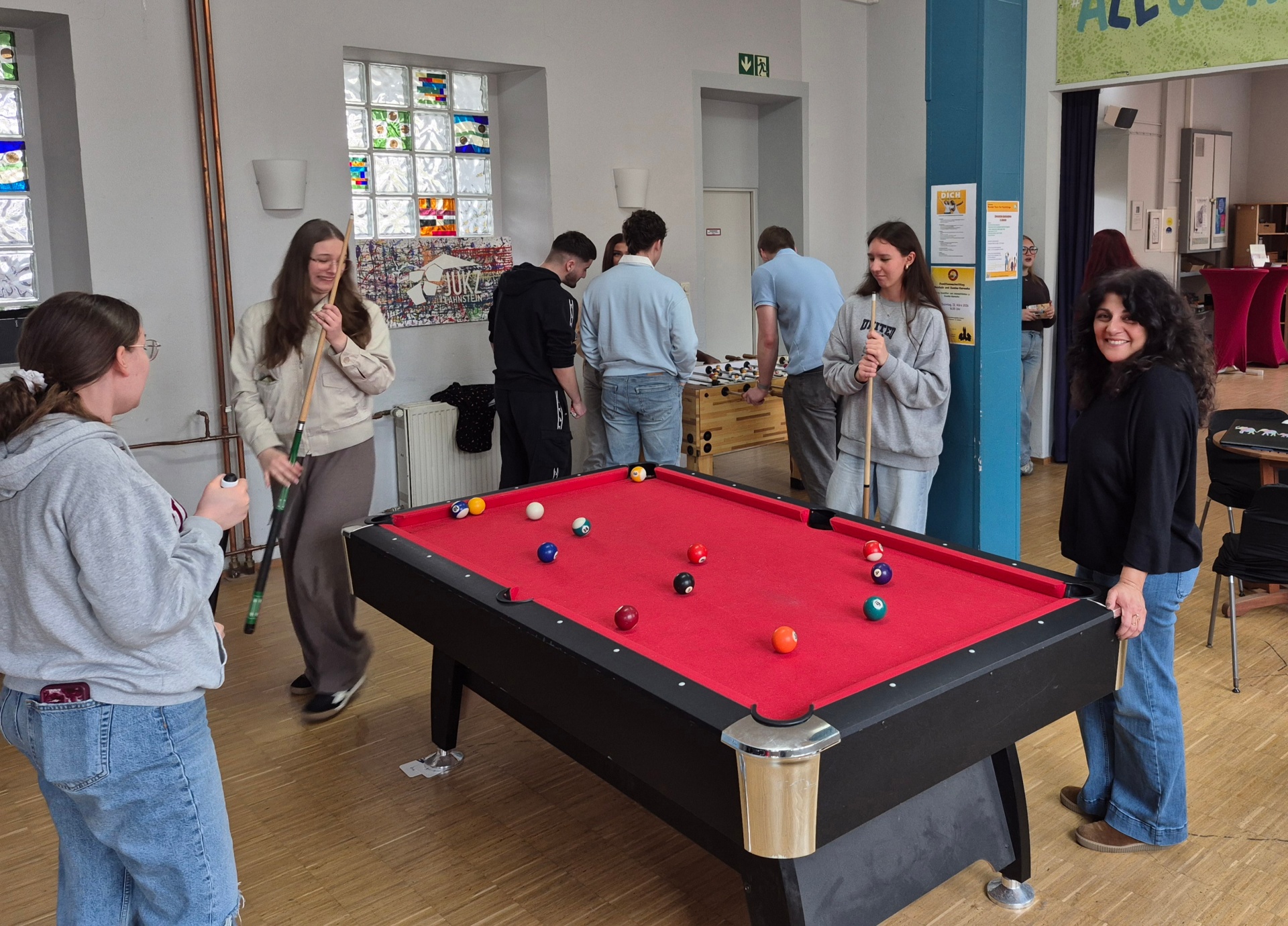 Jugendliche spielen gemeinsam Billard im Jugendkulturzentrum Lahnstein. Rund um den roten Billardtisch stehen mehrere junge Menschen mit Queue in lockerer Atmosphäre, während im Hintergrund weitere Jugendliche kickern und sich unterhalten.