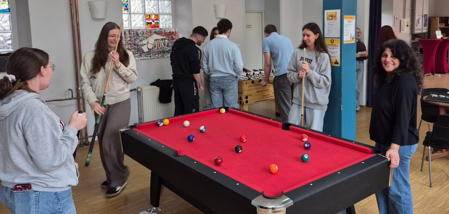 Jugendliche spielen gemeinsam Billard im Jugendkulturzentrum Lahnstein. Rund um den roten Billardtisch stehen mehrere junge Menschen mit Queue in lockerer Atmosphäre, während im Hintergrund weitere Jugendliche kickern und sich unterhalten.