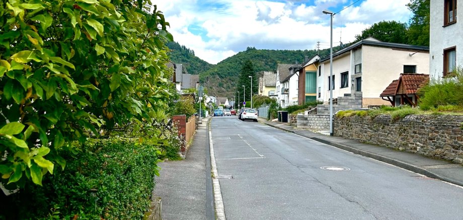 Blick entlang der Straße Hohenrhein in Lahnstein. Die asphaltierte Straße verläuft zwischen Wohnhäusern und begrünten Grundstücken. Links säumen Bäume und Sträucher den Gehweg, im Hintergrund sind bewaldete Hügel unter einem blau-weiß bewölkten Himmel zu sehen.