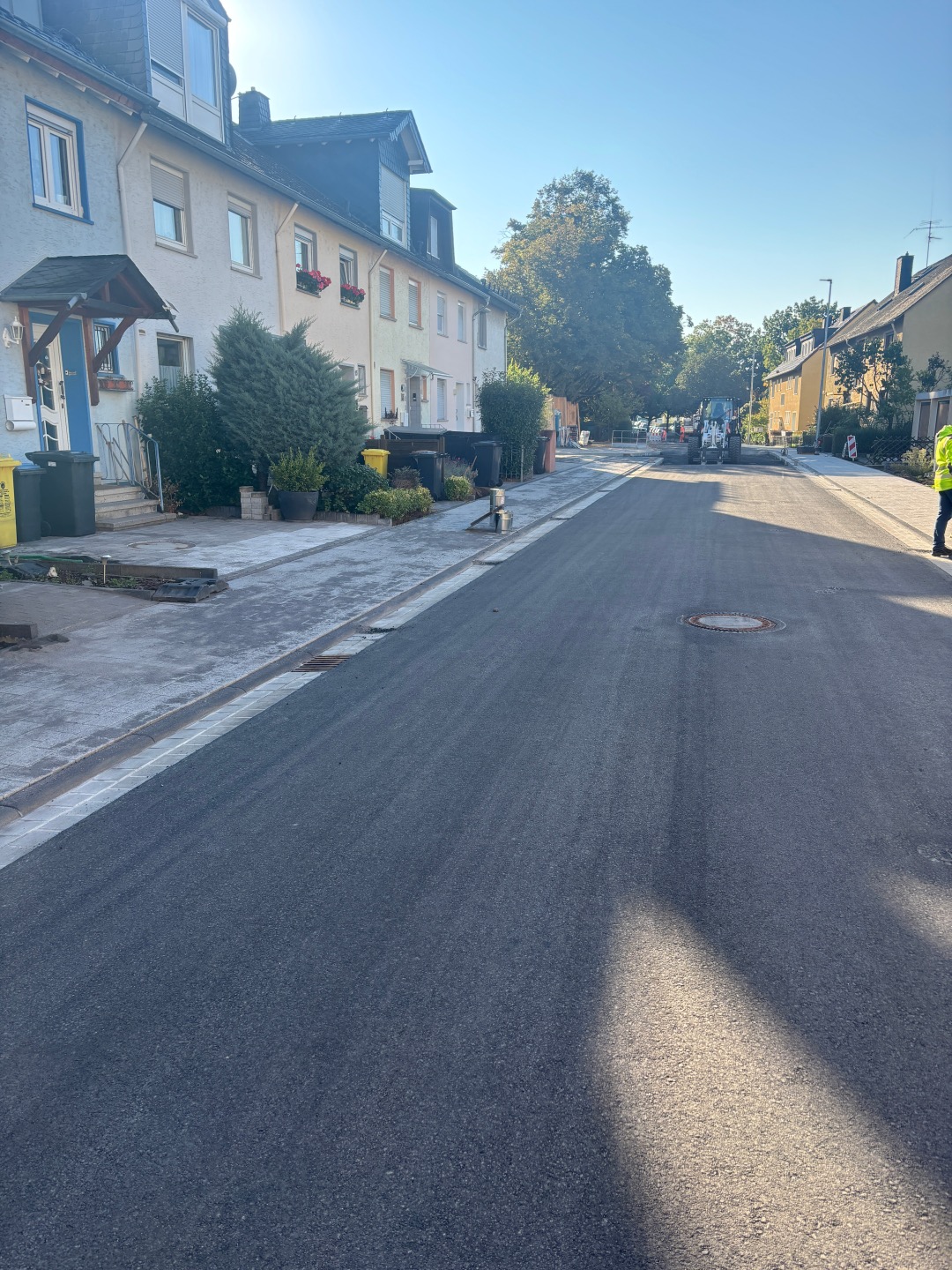 Bllick auf eine Straße, den Lindenweg in Lahnstein, mit frisch asphaltierter Fahrbahn und neu angelegten Gehwegen. Links stehen mehrere Wohnhäuser mit begrünten Vorgärten. Im Hintergrund sind eine Baumaschine und Absperrungen zu sehen. Die Sonne scheint auf den neu gestalteten Straßenzug.