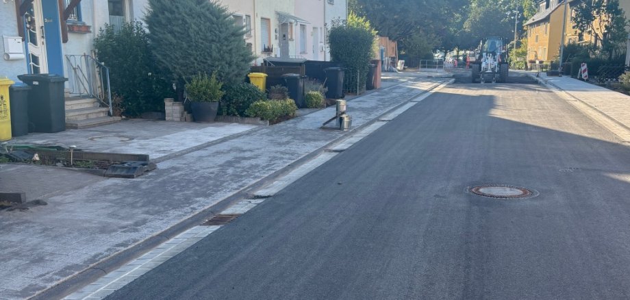 Lindenweg Lahnstein Bllick auf eine Straße, den Lindenweg in Lahnstein, mit frisch asphaltierter Fahrbahn und neu angelegten Gehwegen. Links stehen mehrere Wohnhäuser mit begrünten Vorgärten. Im Hintergrund sind eine Baumaschine und Absperrungen zu sehen. Die Sonne scheint auf den neu gestalteten Straßenzug.