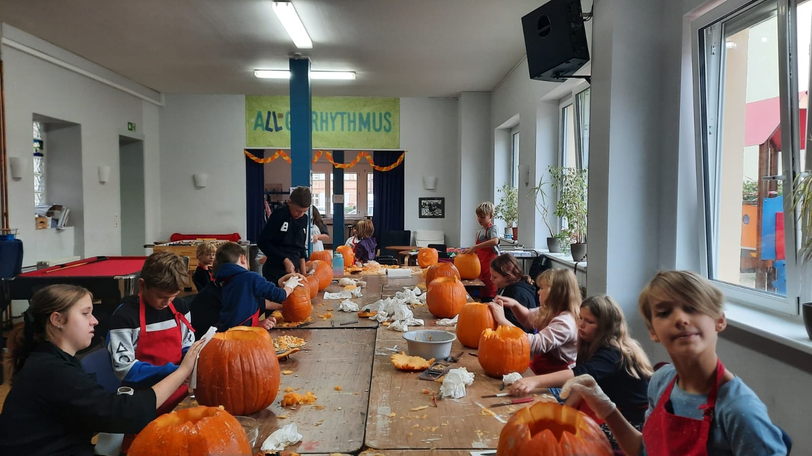 Mehrere Kinder und Jugendliche sitzen und stehen an einem langen Holztisch in einem hellen Raum des Jugendkulturzentrums und schnitzen gemeinsam Kürbisse. Auf dem Tisch stehen zahlreiche große orangefarbene Kürbisse, einige davon bereits ausgehöhlt oder mit eingeschnittenen Gesichtern. Dazwischen liegen Kürbisreste, Werkzeuge, Schüsseln und Papiertücher. Im Hintergrund sind weitere Personen, ein Billardtisch und große Fenster zu sehen.