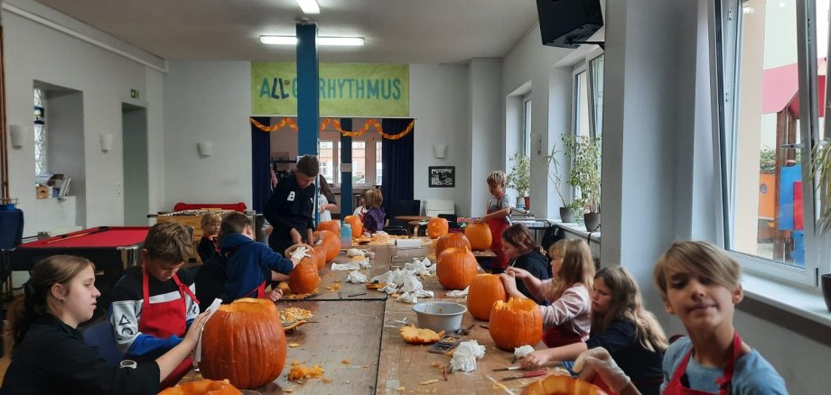 Kürbisschnitzen im Jugendkulturzentrum Lahnstein Mehrere Kinder und Jugendliche sitzen und stehen an einem langen Holztisch in einem hellen Raum des Jugendkulturzentrums und schnitzen gemeinsam Kürbisse. Auf dem Tisch stehen zahlreiche große orangefarbene Kürbisse, einige davon bereits ausgehöhlt oder mit eingeschnittenen Gesichtern. Dazwischen liegen Kürbisreste, Werkzeuge, Schüsseln und Papiertücher. Im Hintergrund sind weitere Personen, ein Billardtisch und große Fenster zu sehen.