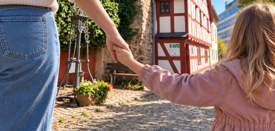 Elternkurs Kess-erziehen Eine erwachsene Person und ein Kind sind von hinten zu sehen und halten sich an den Händen. Im Hintergrund stehen das historische Lahnsteiner Stadtmauerhäuschen mit rot-weißem Fachwerk sowie eine teilweise mit Efeu bewachsene Stadtmauer. Links befinden sich eine historische Wasserpumpe, eine Bank und ein bepflanzter Kübel. Der Weg ist mit Kopfsteinpflaster befestigt.