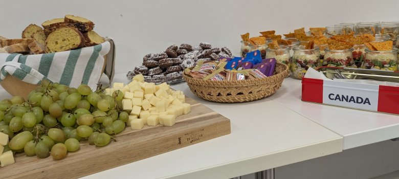 Spieleabend Stadtbücherei Auf einem weißen Tisch steht ein abwechslungsreiches Buffet mit Brot, Weintrauben, Käsewürfeln, Gebäck, Süßigkeiten und kleinen Salaten in Gläsern. Daneben steht eine Serviettenbox mit der Aufschrift „Canada“.