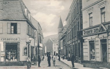 Historische Schwarz-Weiß-Aufnahme einer Straße in Niederlahnstein aus dem Jahr 1907. Auf beiden Seiten stehen dicht aneinandergebaute Wohn- und Geschäftshäuser. Mehrere Erwachsene und Kinder befinden sich auf der Straße und den Gehwegen. In der Straßenmitte verlaufen Straßenbahnschienen, im Hintergrund ist ein Kirchturm zu sehen.